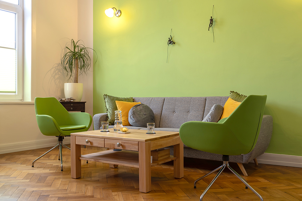 Eine Aufnahme der gemütlichen Praxisräume mit einer grünen Wand einem Holztisch, zwei grünen Sesseln und einem grauen Sofa.
